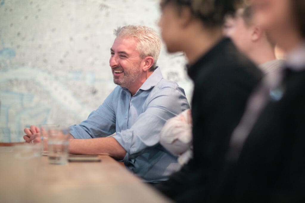 A male sat at a table in a boardroom meeting, with a slightly blurred effect on a female in the foreground.